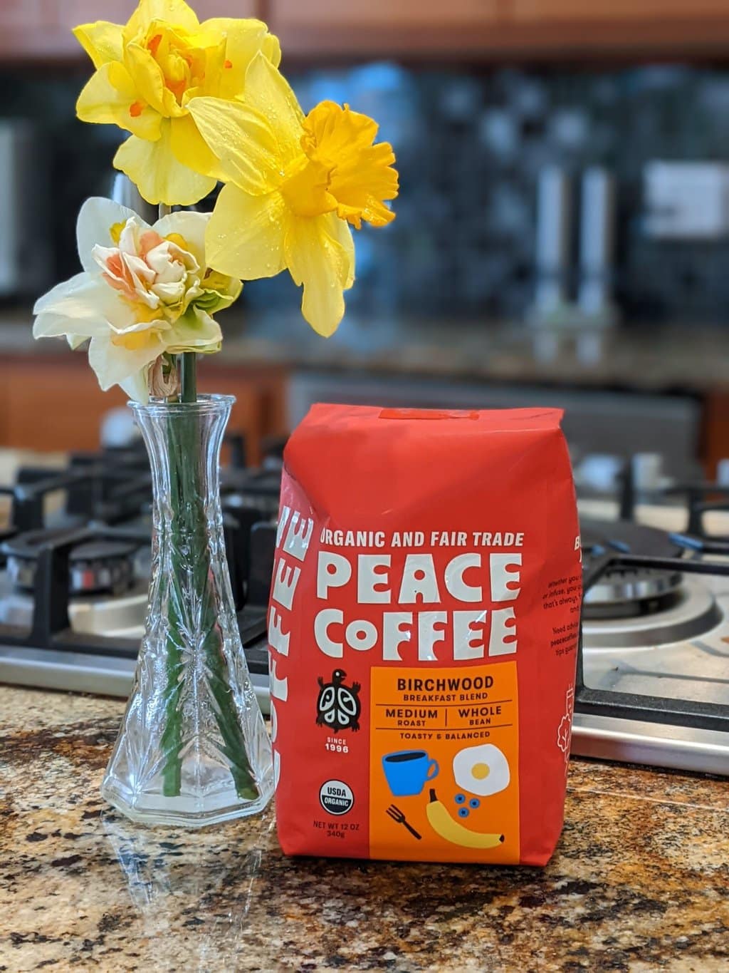 On the kitchen table, next to a vase with daffodils, there is a package of Peace Coffee Birchwood Breakfast Blend Medium Roast.