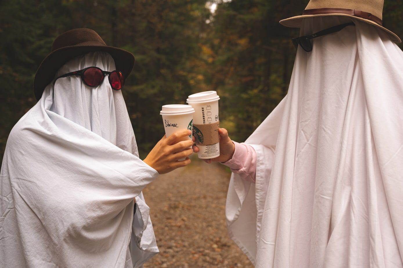 two people in ghost costumes holding white cardboard glasses with Starbucks coffee drink